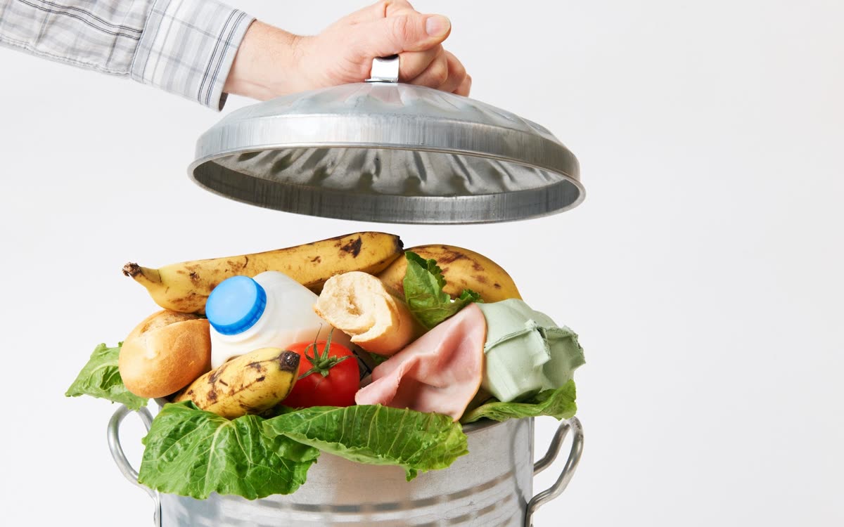 A hand lifting a metal bin lid revealing fresh food being thrown away — bananas, lettuce, bread, milk, a tomato, ham, and an egg carton